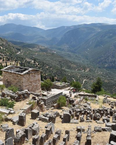 The image shows ancient stone ruins on a mountainside with a view of a valley and distant mountains, featuring a prominent rectangular stone building (the Treasury of the Athenians) and scattered column bases.