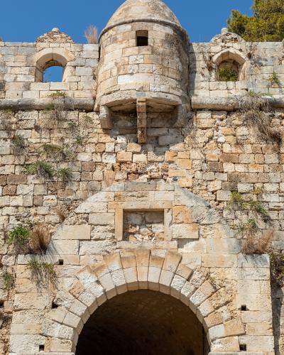 A close-up view of a stone fortress wall featuring a large arched gateway, a central cylindrical tower with a domed roof, and smaller arched windows, set against a clear blue sky.