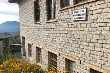 A stone building with wooden window frames and a white sign displaying Greek text "ΡΙΖΑΡΕΙΟΣ ΧΕΙΡΟΤΕΧΝΙΚΗ ΣΧΟΛΗ ΜΟΝΟΔΕΝΔΡΙΟΥ" (Rizarios Handicraft School of Monodendri), set against a backdrop of mountains and yellow flowers.