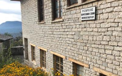 A stone building with wooden window frames and a white sign displaying Greek text "ΡΙΖΑΡΕΙΟΣ ΧΕΙΡΟΤΕΧΝΙΚΗ ΣΧΟΛΗ ΜΟΝΟΔΕΝΔΡΙΟΥ" (Rizarios Handicraft School of Monodendri), set against a backdrop of mountains and yellow flowers.