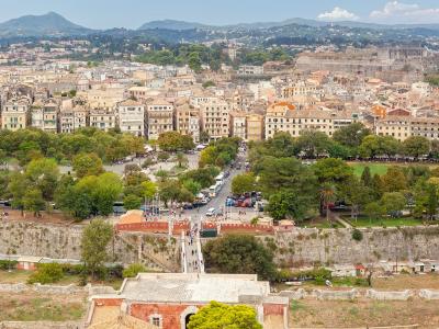 An aerial view of a historic walled city with dense terracotta-roofed buildings, a large park (Spianada), and a prominent fortress on a hill in the background.