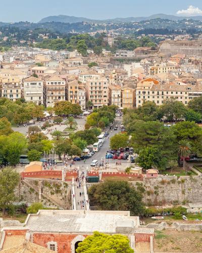 An aerial view of a historic walled city with dense terracotta-roofed buildings, a large park (Spianada), and a prominent fortress on a hill in the background.