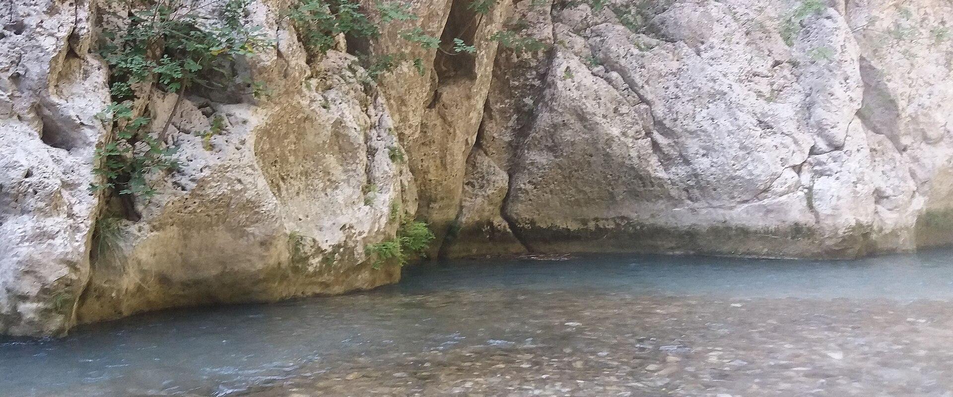 A view of a clear, shallow river flowing at the base of a steep, rocky limestone cliff face with sparse vegetation.