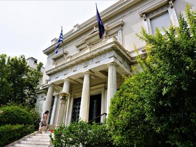 A low-angle shot of a white neoclassical building with a portico of fluted columns, a Greek flag, and a blue flag, with a woman walking up the front steps.