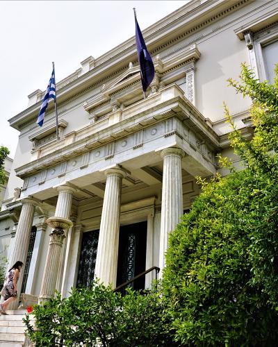 A low-angle shot of a white neoclassical building with a portico of fluted columns, a Greek flag, and a blue flag, with a woman walking up the front steps.