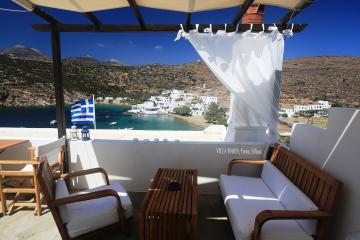 A furnished terrace with wooden furniture and a Greek flag overlooking a coastal village with white buildings and a bay.