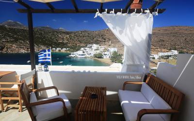 A furnished terrace with wooden furniture and a Greek flag overlooking a coastal village with white buildings and a bay.