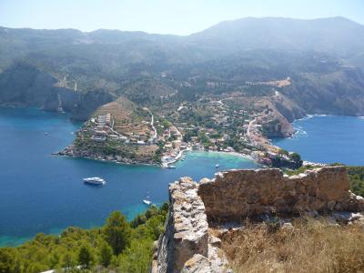 A high-angle view of a coastal village nestled in a cove with a small harbor, surrounded by hills and blue sea, with ancient stone ruins in the immediate foreground.