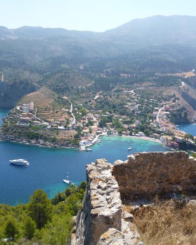 A high-angle view of a coastal village nestled in a cove with a small harbor, surrounded by hills and blue sea, with ancient stone ruins in the immediate foreground.