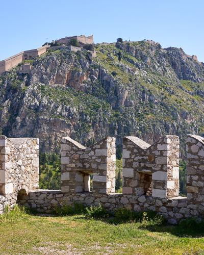 A view from the battlements of a stone fortress looking up a steep, rocky hill towards a larger fortification structure at the summit.
