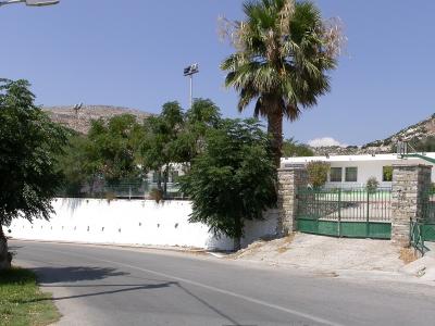 A paved road curves past a white-walled sports court with green fencing and a tall palm tree, set against a backdrop of dry, rocky hills under a clear blue sky.