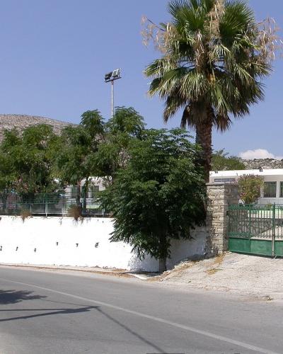 A paved road curves past a white-walled sports court with green fencing and a tall palm tree, set against a backdrop of dry, rocky hills under a clear blue sky.