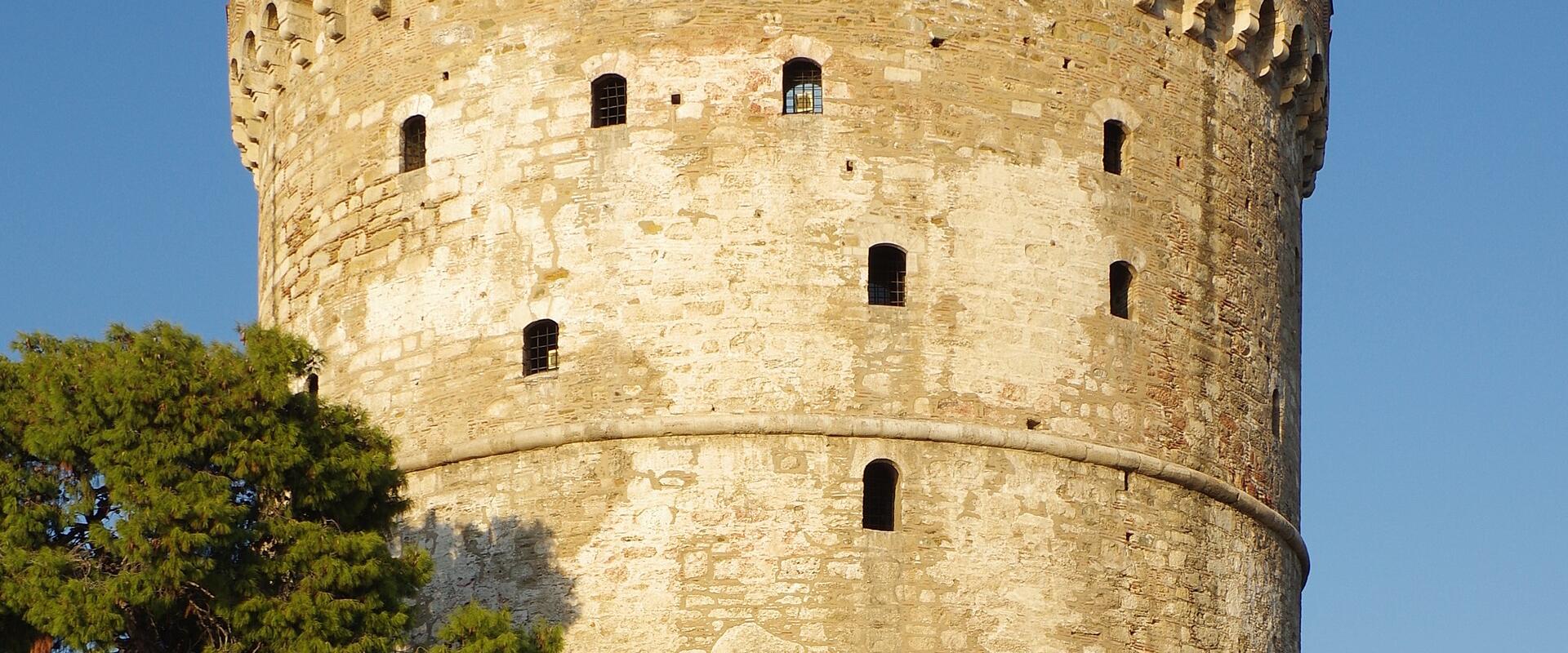 The image shows the White Tower of Thessaloniki, a historic cylindrical stone tower, with a Greek flag flying from its top, set against a clear blue sky, with people relaxing on the grass in the foreground.