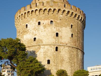 The image shows the White Tower of Thessaloniki, a historic cylindrical stone tower, with a Greek flag flying from its top, set against a clear blue sky, with people relaxing on the grass in the foreground.