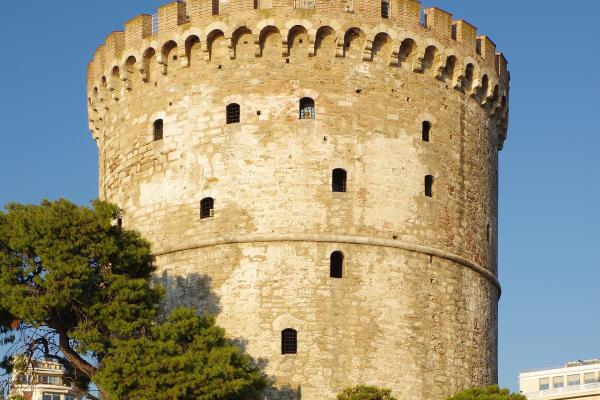 The image shows the White Tower of Thessaloniki, a historic cylindrical stone tower, with a Greek flag flying from its top, set against a clear blue sky, with people relaxing on the grass in the foreground.
