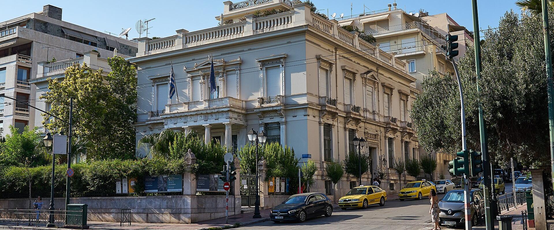 A large, white neoclassical building with a colonnaded entrance, flying Greek and European Union flags, situated on a city street with a sidewalk and trees in the foreground.