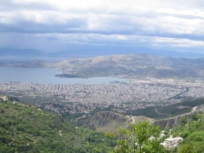 A high-angle panoramic view of a large coastal city (Volos) and its harbor, seen from a green, forested hillside with a cloudy sky above.