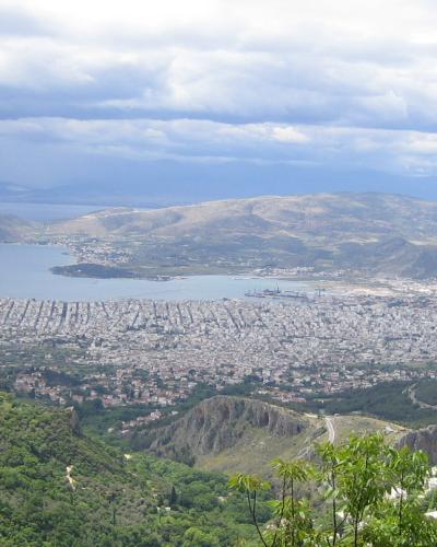 A high-angle panoramic view of a large coastal city (Volos) and its harbor, seen from a green, forested hillside with a cloudy sky above.