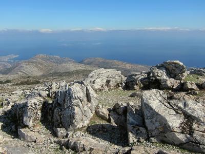 A rocky, arid mountain summit with large boulders in the foreground and a view of the sea and distant islands on the horizon.