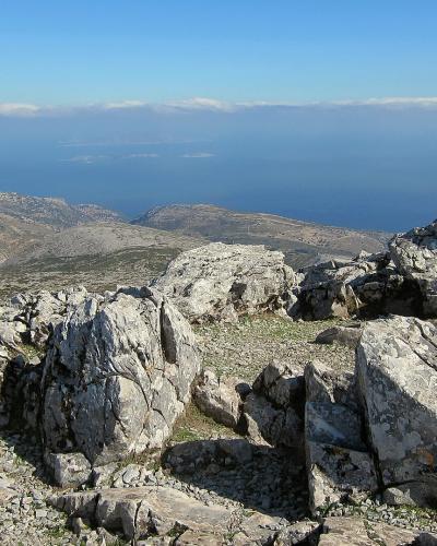 A rocky, arid mountain summit with large boulders in the foreground and a view of the sea and distant islands on the horizon.