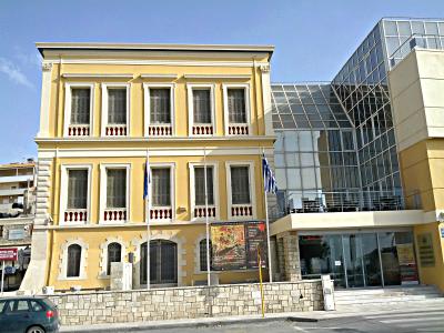 A yellow neoclassical building with a modern glass extension, flying Greek flags, and a poster board in front.
