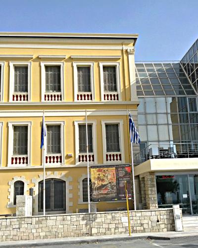 A yellow neoclassical building with a modern glass extension, flying Greek flags, and a poster board in front.