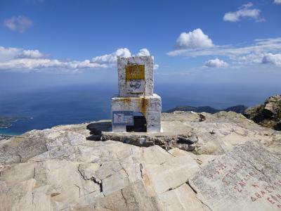 A white stone summit marker with a yellow sign and a blue survey plaque reading "1204" stands on a rocky peak overlooking the sea.
