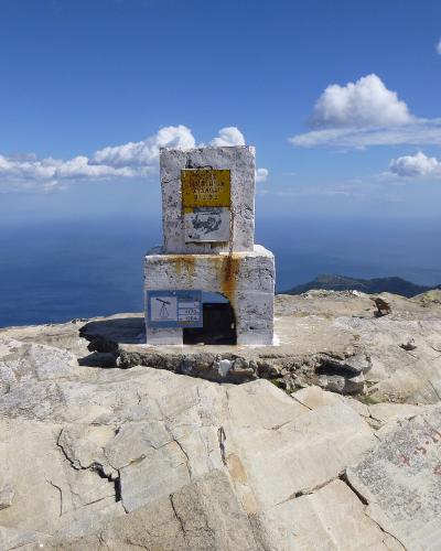 A white stone summit marker with a yellow sign and a blue survey plaque reading "1204" stands on a rocky peak overlooking the sea.
