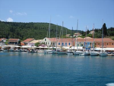 A harbor scene showing a row of colorful neoclassical buildings with terracotta roofs along a waterfront promenade, backed by a green, forested hill, with numerous sailboats and yachts moored in the blue water.