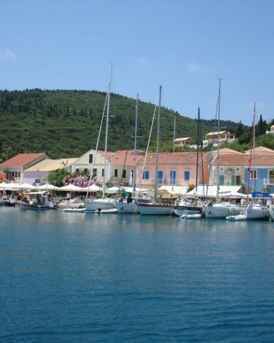 A harbor scene showing a row of colorful neoclassical buildings with terracotta roofs along a waterfront promenade, backed by a green, forested hill, with numerous sailboats and yachts moored in the blue water.