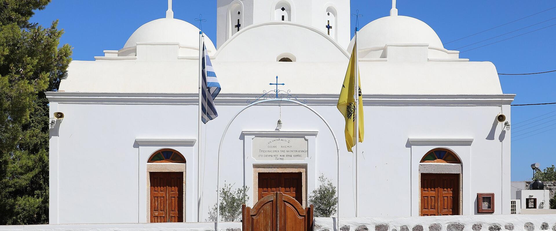 A white-washed Orthodox church with three domes, a stone wall, and Greek and yellow flags, featuring a plaque above the central door.