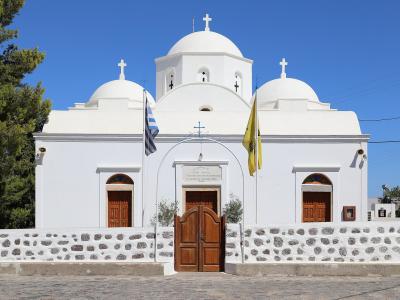 A white-washed Orthodox church with three domes, a stone wall, and Greek and yellow flags, featuring a plaque above the central door.