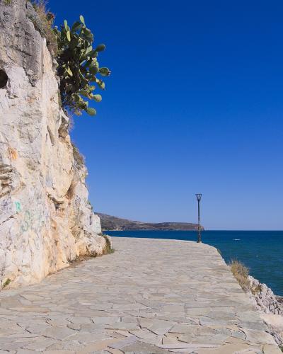 A paved stone coastal promenade running alongside a rocky cliff face with a cactus plant, overlooking the sea under a clear blue sky.