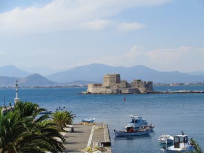 A stone fortress situated on a small islet in the middle of a harbor, with a lighthouse, palm trees, and fishing boats in the foreground and mountains in the background.