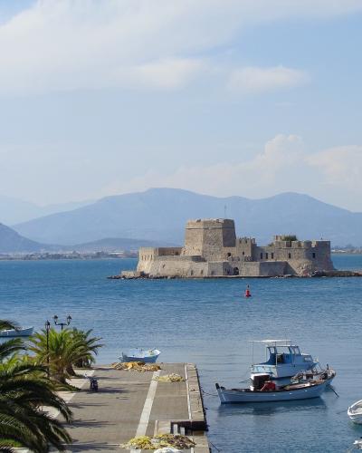 A stone fortress situated on a small islet in the middle of a harbor, with a lighthouse, palm trees, and fishing boats in the foreground and mountains in the background.