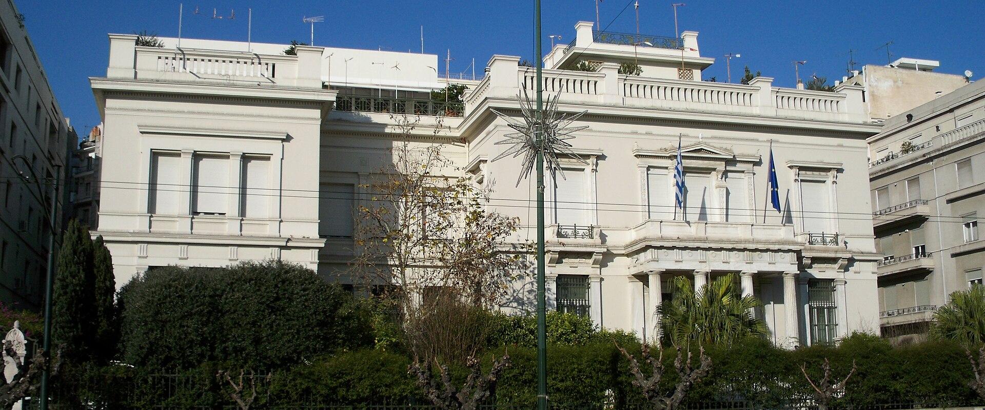 A large, white neoclassical building with a colonnaded entrance and Greek and European Union flags flying in front, situated on a city street with trees and a sidewalk.