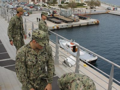 US Navy sailors on the deck of a ship operating a winch, with the harbor and city of Ermoupoli visible in the background.