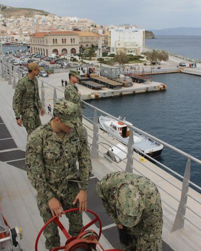 US Navy sailors on the deck of a ship operating a winch, with the harbor and city of Ermoupoli visible in the background.