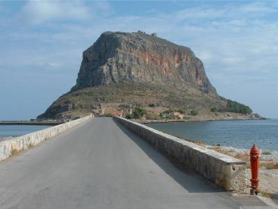 A paved causeway with low stone walls leading towards a large, steep rocky island topped with medieval ruins, with a red fire hydrant visible in the foreground.