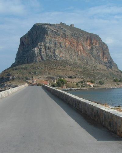 A paved causeway with low stone walls leading towards a large, steep rocky island topped with medieval ruins, with a red fire hydrant visible in the foreground.