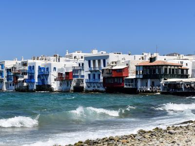 A row of white-washed buildings with colorful balconies and overhangs built directly on the sea edge, with a large cruise ship visible on the far left and a rocky shoreline in the foreground.