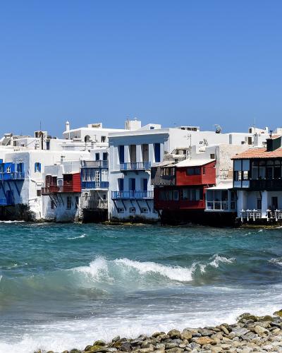 A row of white-washed buildings with colorful balconies and overhangs built directly on the sea edge, with a large cruise ship visible on the far left and a rocky shoreline in the foreground.