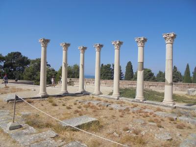 A row of seven standing ancient columns with Corinthian capitals on a terraced archaeological site with dry grass and a blue sky.