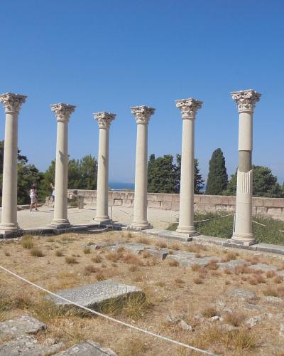 A row of seven standing ancient columns with Corinthian capitals on a terraced archaeological site with dry grass and a blue sky.