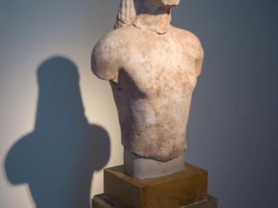 A marble torso of an ancient Greek kouros statue displayed on a pedestal with a museum label in a gallery setting.
