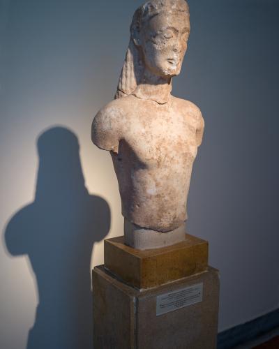 A marble torso of an ancient Greek kouros statue displayed on a pedestal with a museum label in a gallery setting.