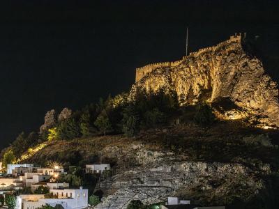 A night photograph showing a fortified acropolis on a rocky hill illuminated by floodlights, overlooking a village with white buildings and stone steps in the foreground.