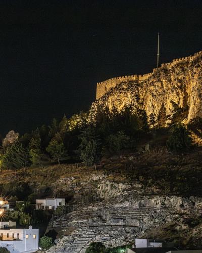 A night photograph showing a fortified acropolis on a rocky hill illuminated by floodlights, overlooking a village with white buildings and stone steps in the foreground.