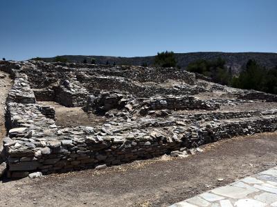 The image shows the stone foundations and ruins of an ancient settlement under a clear blue sky, with a paved walkway in the foreground and hills in the background.