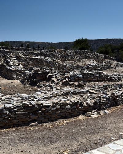 The image shows the stone foundations and ruins of an ancient settlement under a clear blue sky, with a paved walkway in the foreground and hills in the background.